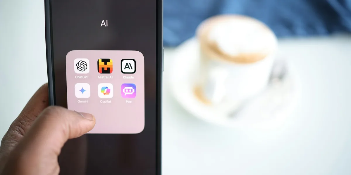 GeminiやClaudeなどのAIアシスタントアプリのフォルダが表示されたスマートフォンと、コーヒーカップ