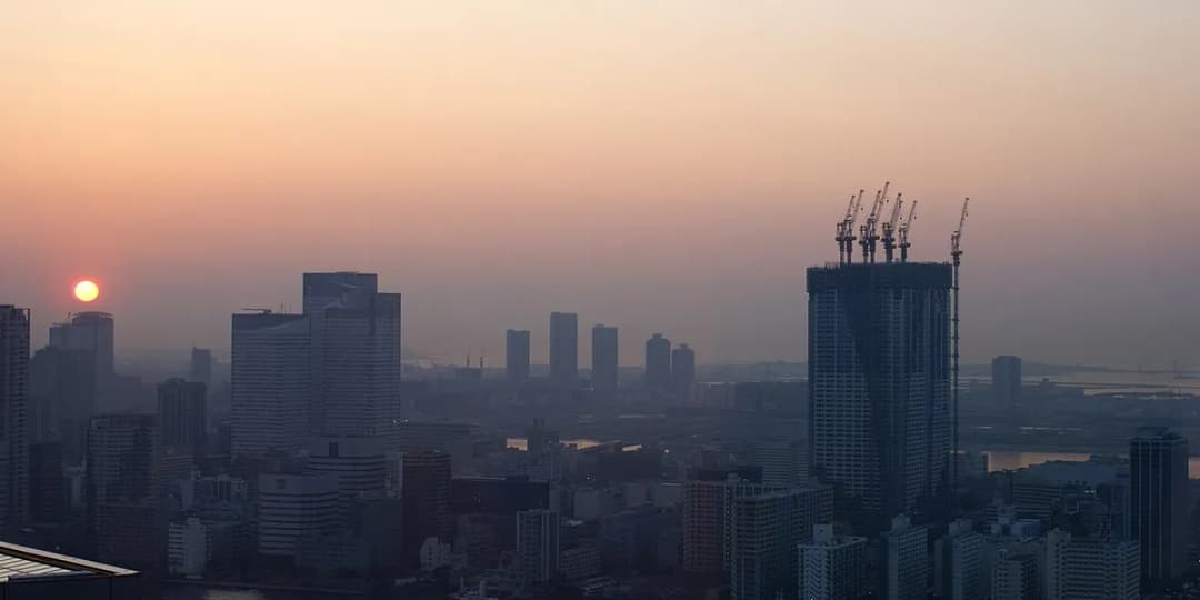 The sun rising through haze over the Tokyo skyline, with cranes on a tower still under construction